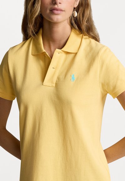 Geltonas medvilninis polo marškinėliai su tekstūruotu paviršiumi, turi apykaklę, tris mygtukus ir šviesiai mėlyną siuvinėtą logotipą ant krūtinės.