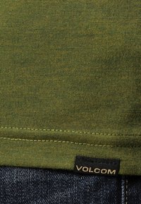 Grøn bomulds T-shirt med et tekstureret strikkemønster, der har en syet kant og et sort "VOLCOM"-mærke på siden.