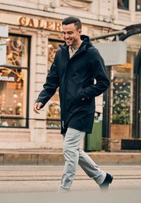 Zwarte hoodie-jack met een relaxte pasvorm, voorzien van een hoge kraag en voorzakken, gecombineerd met lichtgrijze geruite broek en zwarte schoenen.