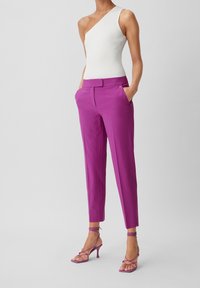 Pantalon violet ajusté de coupe slim avec poches, associé à un top asymétrique blanc côtelé et des talons violets à brides.