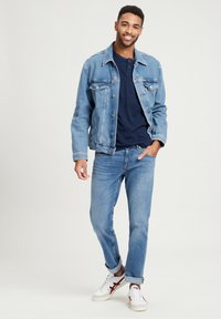 Jeansjack in lichtblauw, gelaagd over een donker marineblauw shirt, gecombineerd met lichtblauwe jeans en witte sneakers met rode accenten.