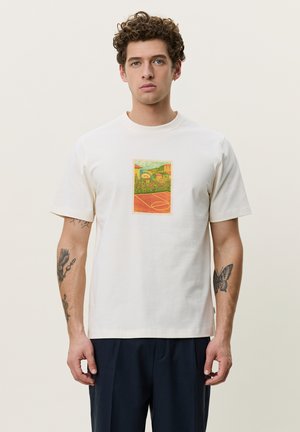 Νεαρός με σγουρά μαλλιά που φοράει λευκό T-shirt με εκτύπωση γηπέδου μπάσκετ και τοπίου, στέκεται μπροστά σε απλό φωτεινό φόντο.