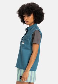 Tyrkis ärmlös skjorta med krage, knäppning framtill och två bröstfickor; bärs över en grå kortärmad skjorta. Modellen har även på sig randiga shorts.