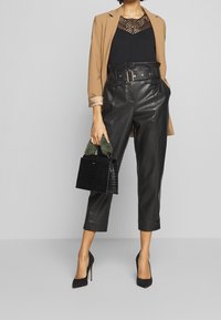 Kahotový blazer přes černý krajkový top, černé kožené zkrácené kalhoty a černé podpatky. Drží malou černou kabelku s krokodýlím vzorem a zelenými akcenty.