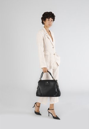 Czarna skórzana torebka z uchwytami zdobionymi łańcuszkiem, teksturowana powierzchnia. Modelka w kremowym garniturze w paski i czarnych szpilkach, stojąca na szarym tle.