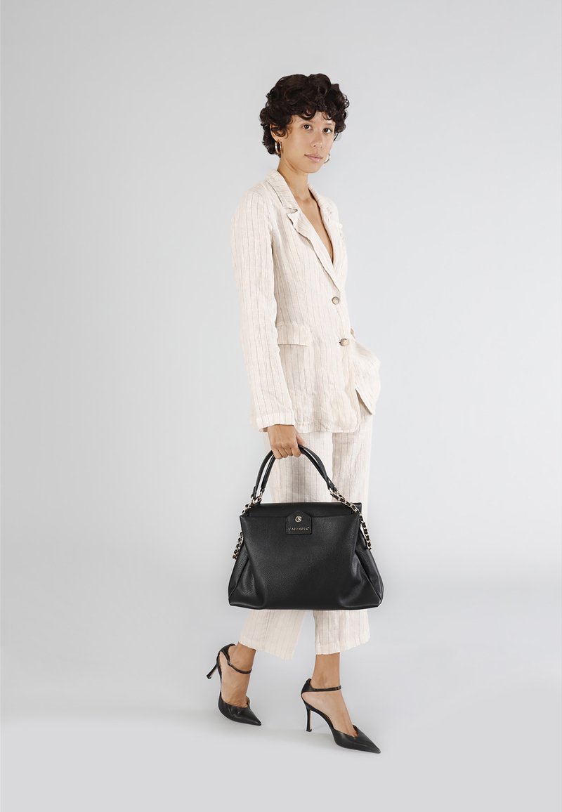 Borsa nera in pelle con manici accentuati da catena e superficie testurizzata. Modello in un completo a righe crema e tacchi a punta neri, in piedi su uno sfondo grigio.