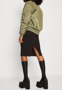 Olivově zelená bombera s ribbed manžetami a lemem, v kombinaci s černou midi sukní s bočním rozparkem a černými koženými vysokými botami na bloku.