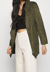 Olivgrön mockajacka med dragkedja framtill, huva och sidofickor; slät textur och enkel design. Bärs över en svart topp och ljusa byxor.