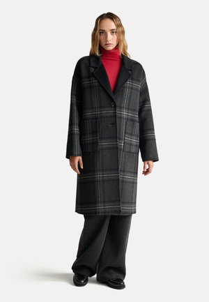 Cappotto di lana a quadri in tonalità di grigio con dettagli neri, chiusura a un bottone e due tasche frontali; indossato su un dolcevita rosso.