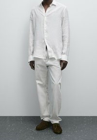 Homme portant une chemise à rayures blanches boutonnée, un jean gris clair et des chaussures à lacets en daim brun foncé, debout sur un sol gris moucheté.