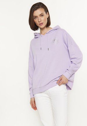 Jonge vrouw draagt een lichtpaarse oversized hoodie met een klein bloemmotief en een witte broek, staand tegen een effen achtergrond.