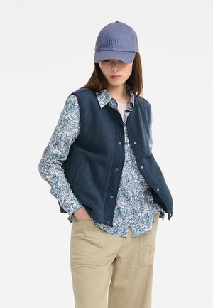 Un gilet en polaire bleu marine sur une chemise à fleurs bleues, avec une casquette bleu clair. La tenue comporte des boutons, des poches et un pantalon beige.
