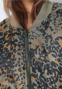 Bomberka w kamuflażu z suwakowym zapięciem z przodu, elastycznymi mankietami i dekoltem. Teksturowany materiał w zielono-czarnych i pomarańczowych wzorach.