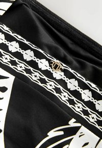 Tissu noir avec des motifs blancs et dorés, présentant des détails complexes et un logo en métal doré. Texture douce et finition lisse.