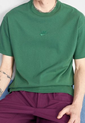 Osoba ma na sobie zieloną koszulkę Nike, fioletowe spodnie, srebrny łańcuszek na szyi i tatuaż na lewym przedramieniu, siedzi w pomieszczeniu.