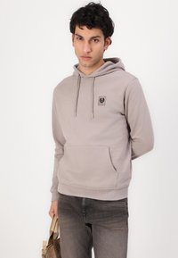 Ανοιχτό γκρι φούτερ με μπροστινή τσέπη, ρυθμιζόμενα κορδόνια και τετράγωνη πατίνα λογότυπου. Συνδυασμένο με σκούρο γκρι τζιν και καφέ τσάντα.