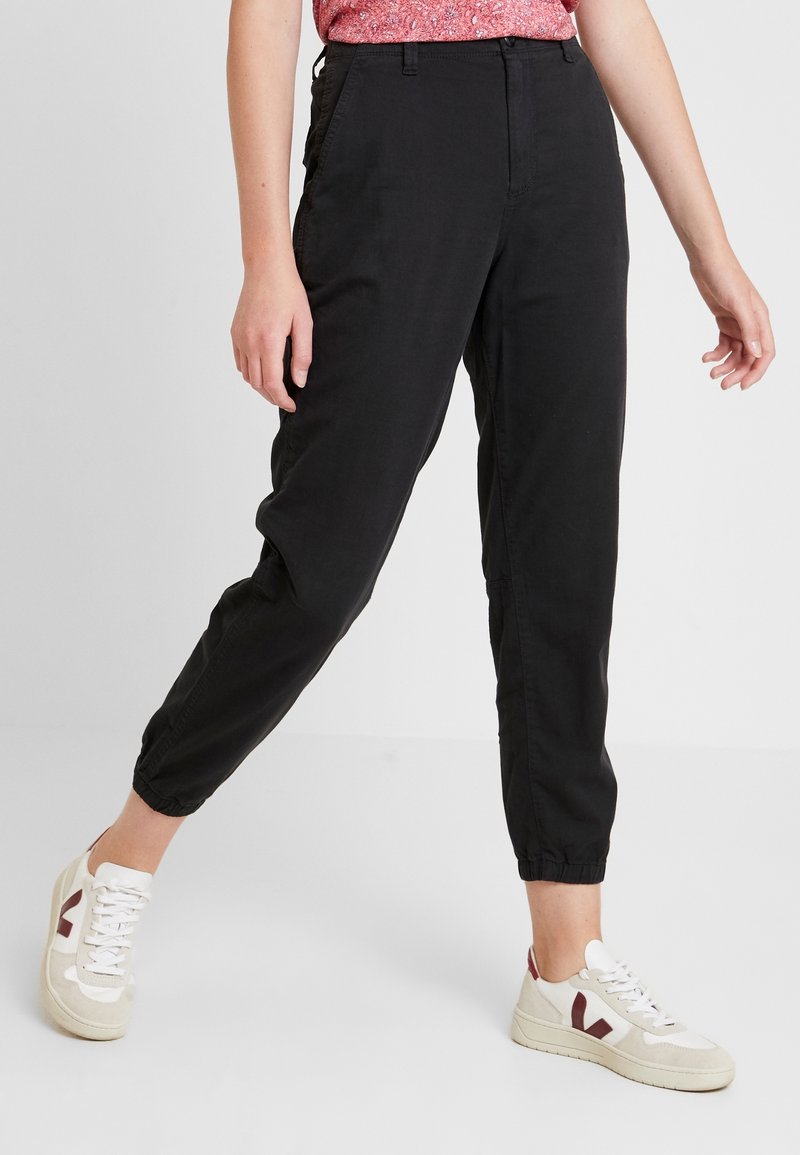 Pantalons de jogging noirs fuselés avec des poignets élastiques, poches latérales et une texture lisse, associés à des baskets blanches et bordeaux.