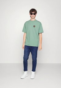 T-shirt verde de algodão com mangas curtas e um emblema quadrado, combinado com jeans azuis e ténis brancos, sobre um fundo neutro.