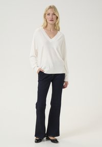 Balts V-veida džemperis ar ribotu apkakli, kombinēts ar tumši ziliem platiem džinsiem un melniem dzīvokļiem. Mīksta struktūra, brīva piegrieztne un mūsdienīgs dizains.