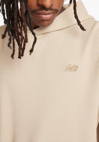 Beige hooded sweatshirt made of ribbed texture fabric, featuring a small embroidered logo on the chest, with a relaxed fit and V-neck design.