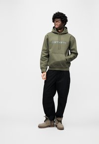 Olivgrüner Kapuzenpullover mit blauem Logo, schwarze Cargohose und beige Stiefeletten mit Schnürsenkeln und Gummisohlen. Glatte Stoffstruktur.