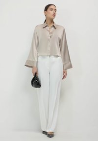 Donna che indossa una blusa lunga a maniche di raso beige, pantaloni bianchi a gamba larga, scarpe nere a punta, mentre tiene una piccola borsa nera.
