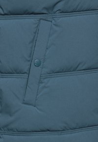 Mörkgrön puffjacka i närbild, med en tryckknapp och väldefinierad sömnad. Materialet ser slätt ut med en något glänsande yta.