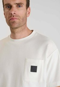 Uomo con capelli corti castani e barba, che indossa una maglietta bianca semplice con un'etichetta quadrata nera sulla tasca del petto.