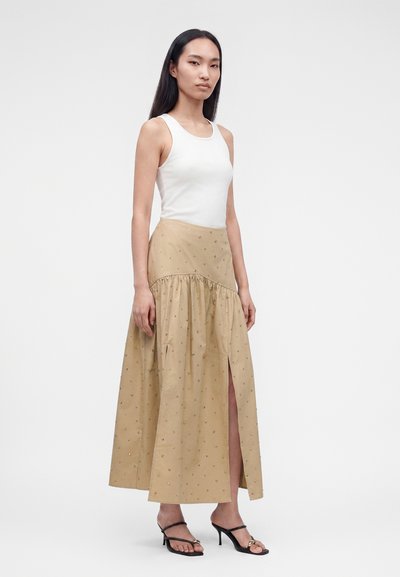 Γυναίκα με λευκό αμάνικο τοπ και μπεζ φούστα μέχρι τους αστραγάλους με σκίσιμο, συνδυασμένο με μαύρα σανδάλια με τακούνι, που στέκεται μπροστά από ένα απλό φόντο.