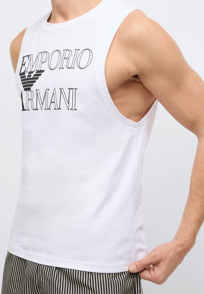 Biała bawełniana koszulka na ramiączkach z czarnym logo „Emporio Armani”, bez rękawów i o luźnym kroju. Ozdobiona widocznym wycięciem pod pachą.