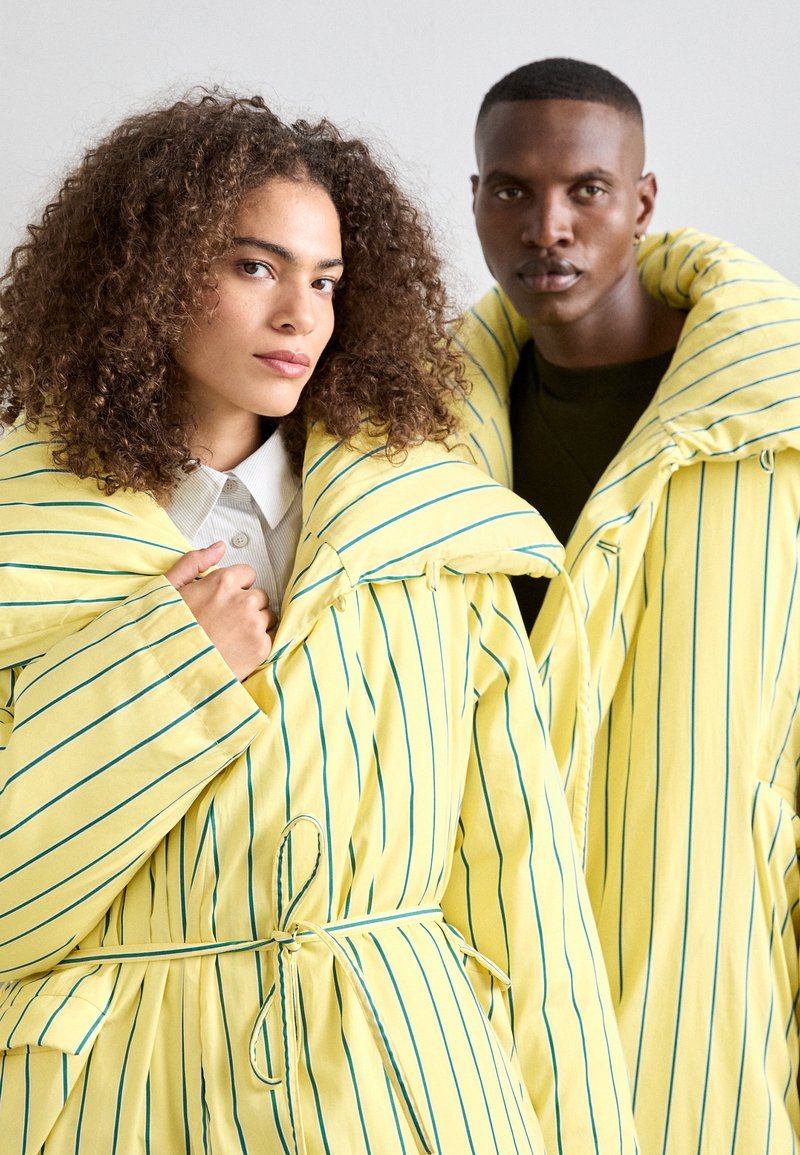 Gelbe gestreifte Oberbekleidung mit einem breiten Kragen, featuring einer taillierten Form und einem weichen Material. Zwei Models präsentieren das Kleidungsstück gemeinsam.