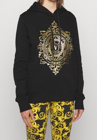 Felpa nera con cappuccio e stampa ornamentale dorata sul davanti, dotata di tasca a marsupio e polsini a coste. Abbinata a leggins gialli con fantasia.