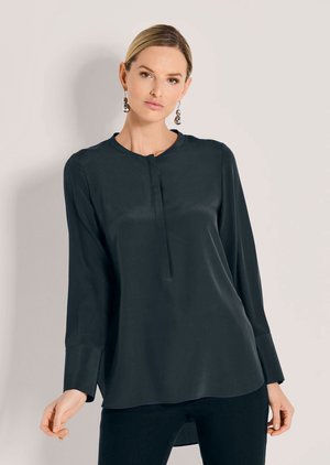 Vrouw met licht haar naar achteren draagt een zwarte blouse met lange mouwen met verborgen knopen aan de voorkant en hangende zilveren oorbellen, poserend tegen een neutrale muur.