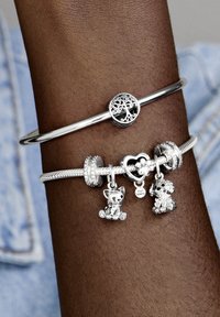 Brazaletes de plata en una muñeca, uno presenta un diseño de árbol, el otro tiene tres charms: un gato, un perro y un corazón con la inscripción "Fur Mom".