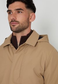 Beige waterdichte jas met een puntkraag, bruine binnenkant en een ritssluiting. Gladde textuur met een lichte glans en zichtbare stiksels.