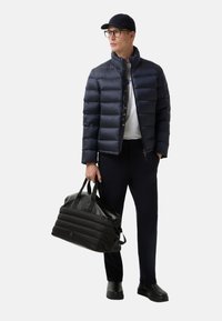 Muž v černé kšiltovce, námořnické péřové bundě, námořnických kalhotách a černých botách, držící černou prošívanou tašku, dívající se do strany na bílém pozadí.