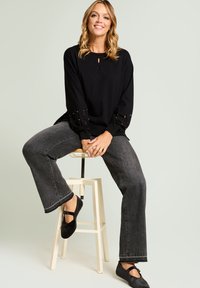 Jersey negro de punto con detalle de abertura en forma de ojo de cerradura, mangas adornadas, combinado con jeans grises acampanados y zapatos planos negros, sentado en un taburete.