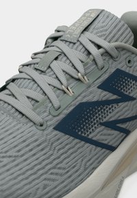 Lähikuva harmaasta juoksukengästä, jossa on teksturoitu kangas, vaaleanharmaat nauhat ja tummansininen logo sivulla, sekä pehmustetun pohjan yksityiskohta.