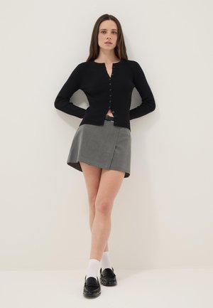 Frau steht vor einer weißen Wand und trägt eine schwarze, geknöpfte Strickjacke, einen grauen asymmetrischen Minirock, weiße Socken und schwarze Loafers.