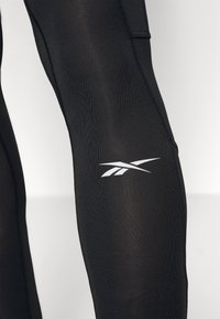 Leggings pretas feitos de material elástico, com um design ajustado, costuras planas e um logótipo branco na coxa.