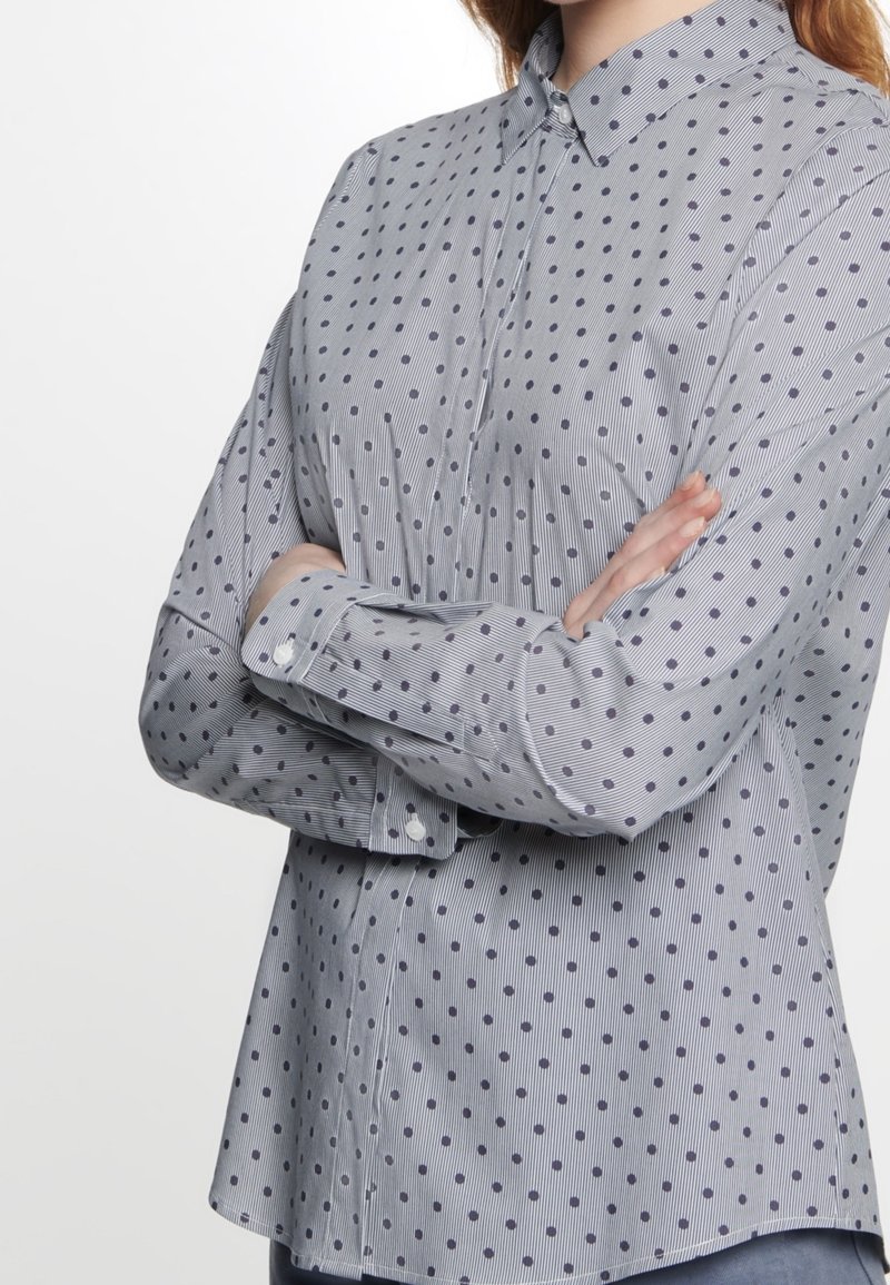 Personne portant une chemise à manches longues boutonnée avec des fines rayures et des pois foncés, bras croisés sur la poitrine.