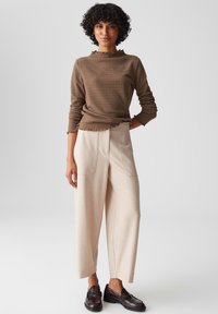 Donna in piedi, che indossa un top lungo a maniche con texture marrone, pantaloni beige a gamba larga con tasche anteriori e mocassini marrone scuro su uno sfondo neutro.