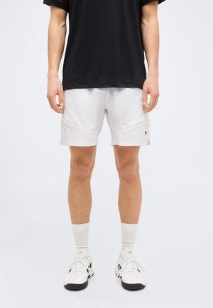 Άνδρας που φοράει μαύρο πουκάμισο, λευκά αθλητικά σορτς, λευκές κάλτσες και λευκά αθλητικά παπούτσια, στέκεται πάνω σε απλό φωτεινό φόντο.