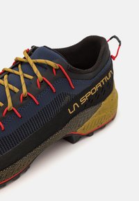 Športová obuv s tmavomodrým sieťovaným zvrškom, žltými šnúrkami, čiernymi akcentmi a robustnou žlto-čierne vzorovanou podrážkou s červenými detailmi.