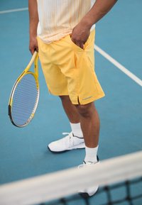Gelbe Shorts mit seitlichen Taschen, kombiniert mit einem weiß-orange gestreiften Shirt, während eine Person auf einem blauen Tennisplatz einen Tennisschläger hält.