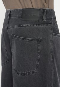 Šedé džíny z denimu s hladkou texturou, vybavené černou koženou nášivkou na pasu a dvěma zadními kapsami. Viditelné jednoduché švy.