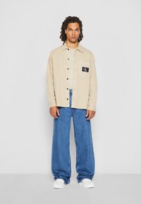 Bēšas pogām apģērbtas krekls ar melnu kabatu, apvienots ar brīviem zilas džinsa biksēm un baltām kedām. Vienkāršs, ikdienišķs dizains, gluda tekstūra.