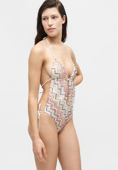 Νέα γυναίκα φοράει πολύχρωμο ολόσωμο μαγιό με σχέδιο ζιγκ-ζαγκ και φιόγκους στο πλάι και μπροστά, στέκεται μπροστά σε απλό φωτεινό φόντο.
