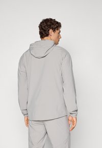 Lysegrå hættejakke med elastiske manchetter og et strømlinet design, lavet af et glat, letvægtsstof. Har en rygdetalje.