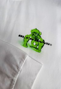 Weißes Baumwoll-T-Shirt mit einem grünen Grafikdesign eines Bären und dem Text "BLCK BNNS." Das Design umfasst feine Details und eine glatte Textur.